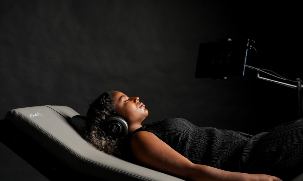 A woman lies on a recliner wearing headphones and looking at a tablet.