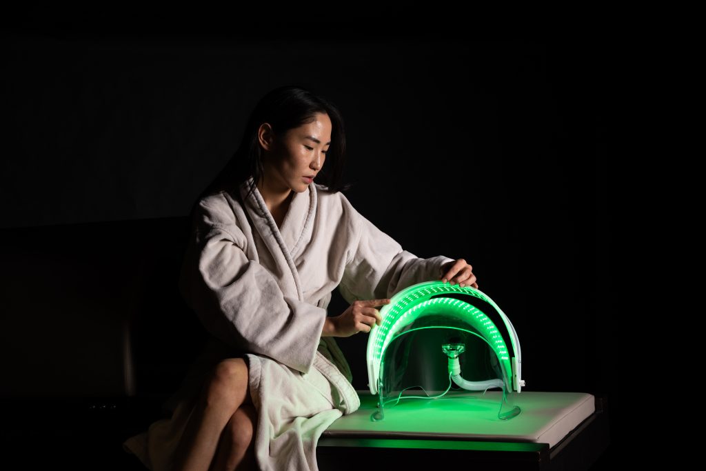 A woman in a white robe sitting next to a green light therapy device.