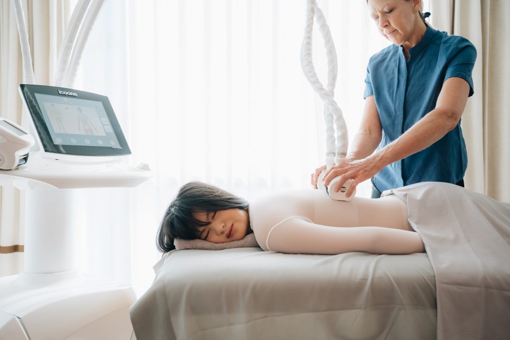 A woman lies on a treatment bed while a therapist uses a device on her back.